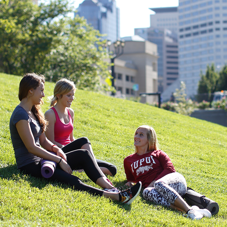Three students sitting on a grassy hill after a run talking with each other.
